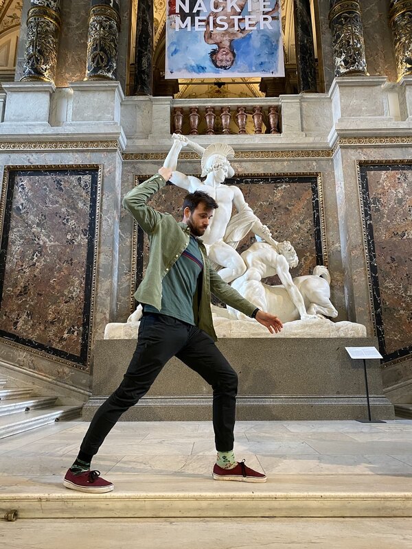 Me in a museum in front of a statue, in an effort to imitate the statue's pose. I have my legs stretched forwards, one arm straight in front of me and my other arm over me as if I were holding a dagger.