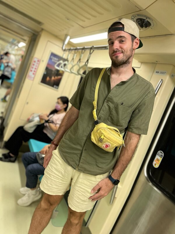 Me standing in a train compartment. I wear a snapback cap backwards, a olive shirt with a yellow hip pack, diagonally around the waist and khaki shorts. I smile tiredly towards the camera.