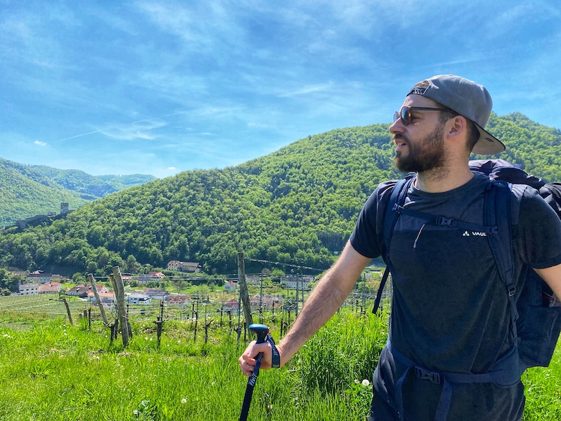 Niq wearing sunglasses and a snapback cap in his hiking gear looking into the distance with a lush green mountain and a vineyard in the background