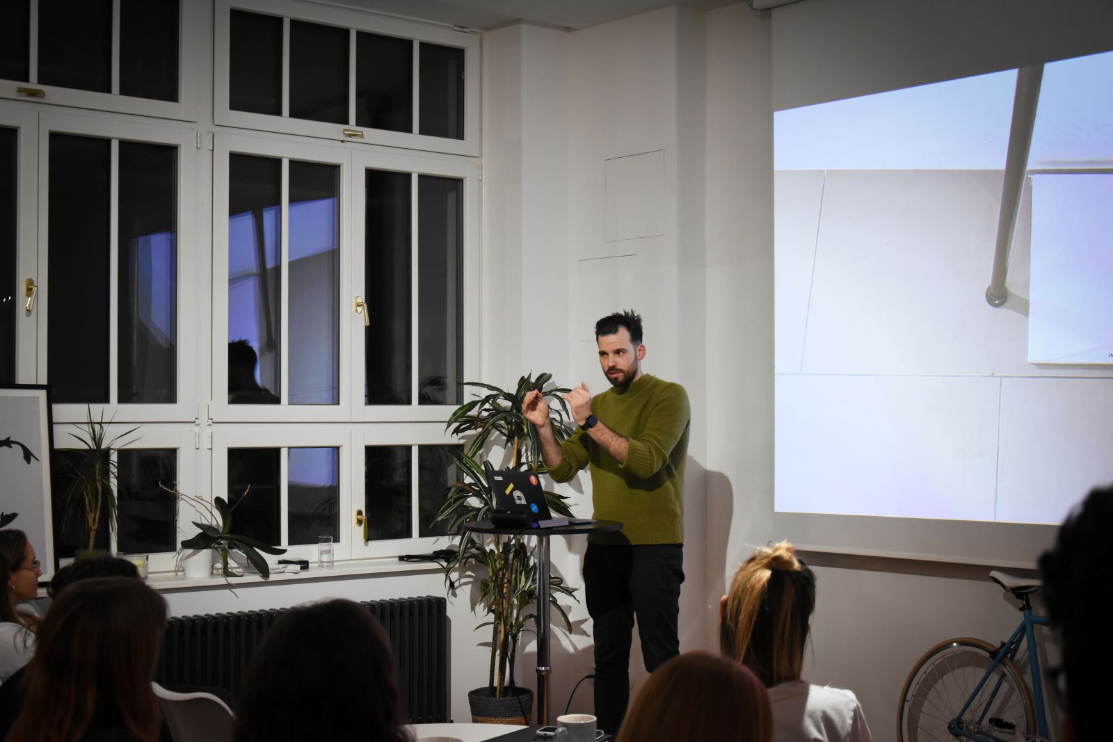 Niq wearing a green woolen sweater next to an indoor plant. He's on stage in front of a crowd gesturing next to the projector screen