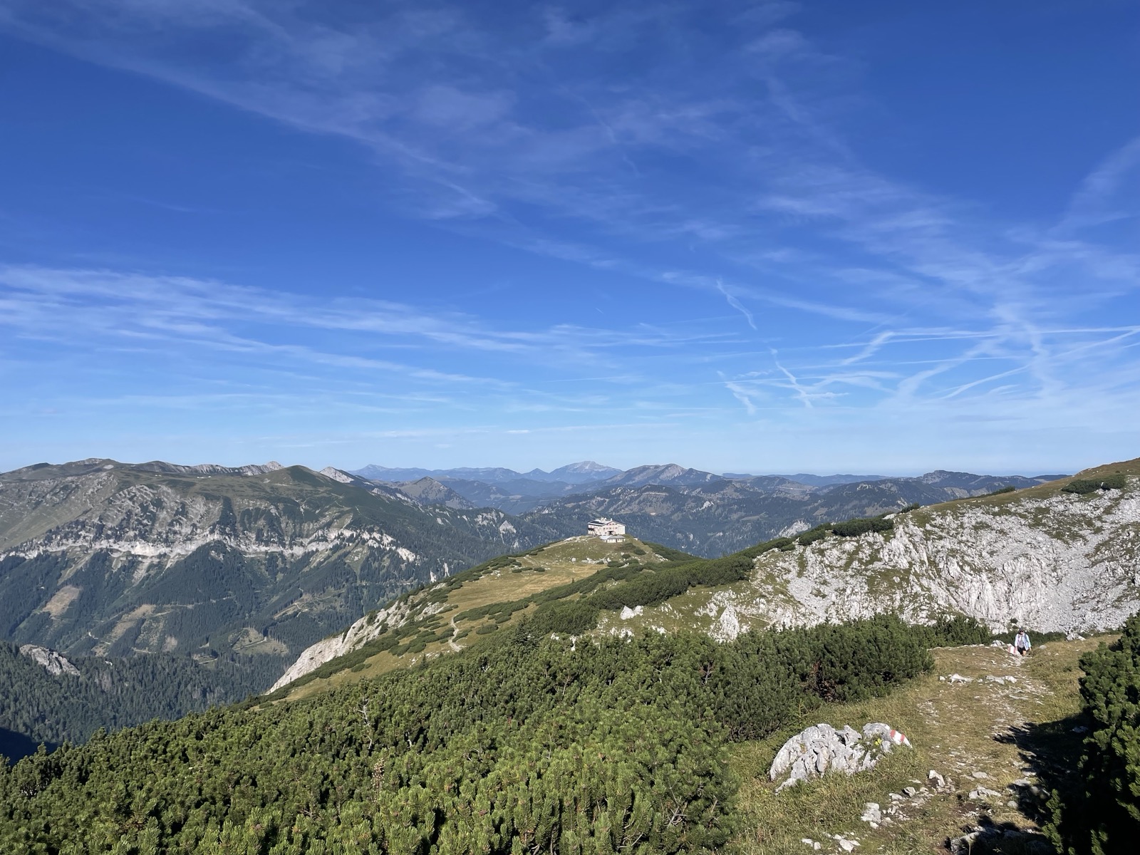 "A mountain landscape, with a blue sky and some scattered clouds above it. At the end of the mountain ridge lies a cabin."