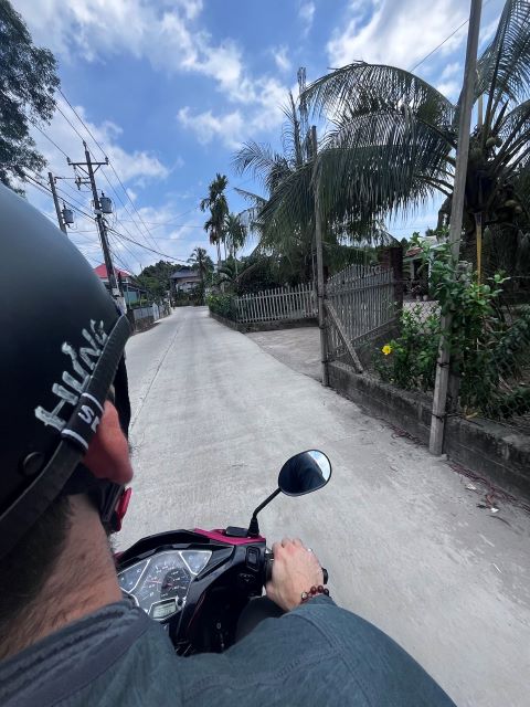 "Me riding a scooter on a street in Phu Quoc. I'm wearing a black helmet and you can see me from the backseat's perspective. The sky is blue with shattered clouds and the vegetation is filled with palm trees on the side of the street."